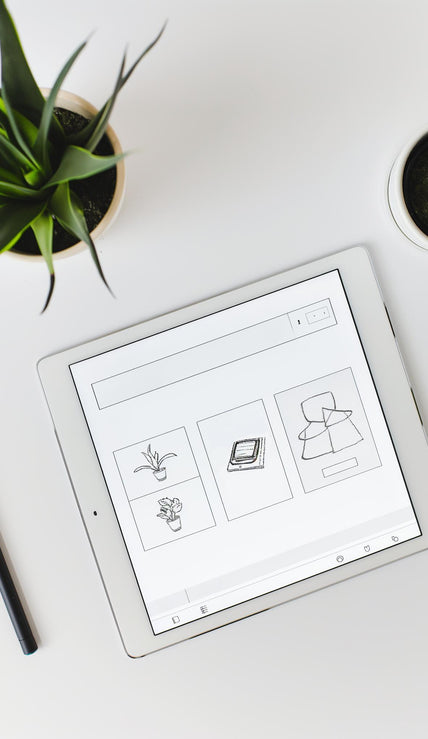 Example product is shown on a tablet with simple sketches of plants and objects, set on a white desk beside two potted succulents, a black pen, and a white stylus.