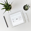 Example product is shown on a tablet with simple sketches of plants and objects, set on a white desk beside two potted succulents, a black pen, and a white stylus.